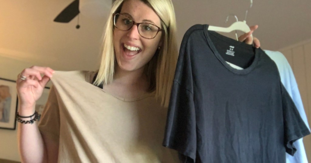 woman smiling and wearing a tan oversized t-shirt holding other t-shirts