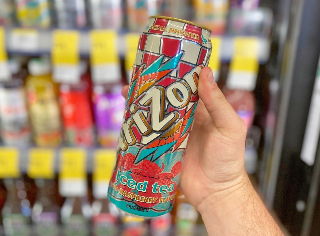 person holding up a can of arizona iced tea in front of refrigerated store case of beverages
