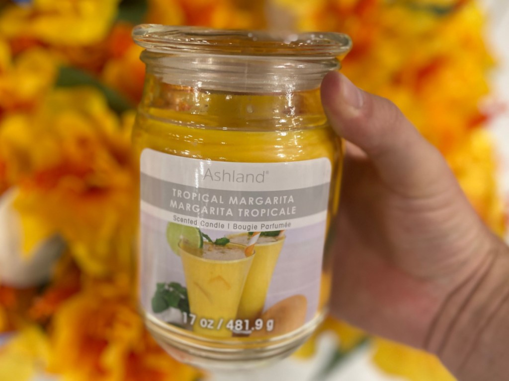 hand holding tropical margarita scented jar candle in front of yellow and orange flowers