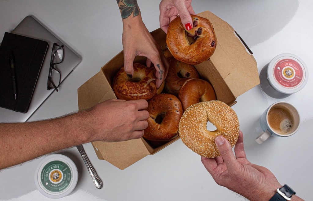four hands reaching for bagels out of the Einstein Bros Bagels box with two tubs of shmear
