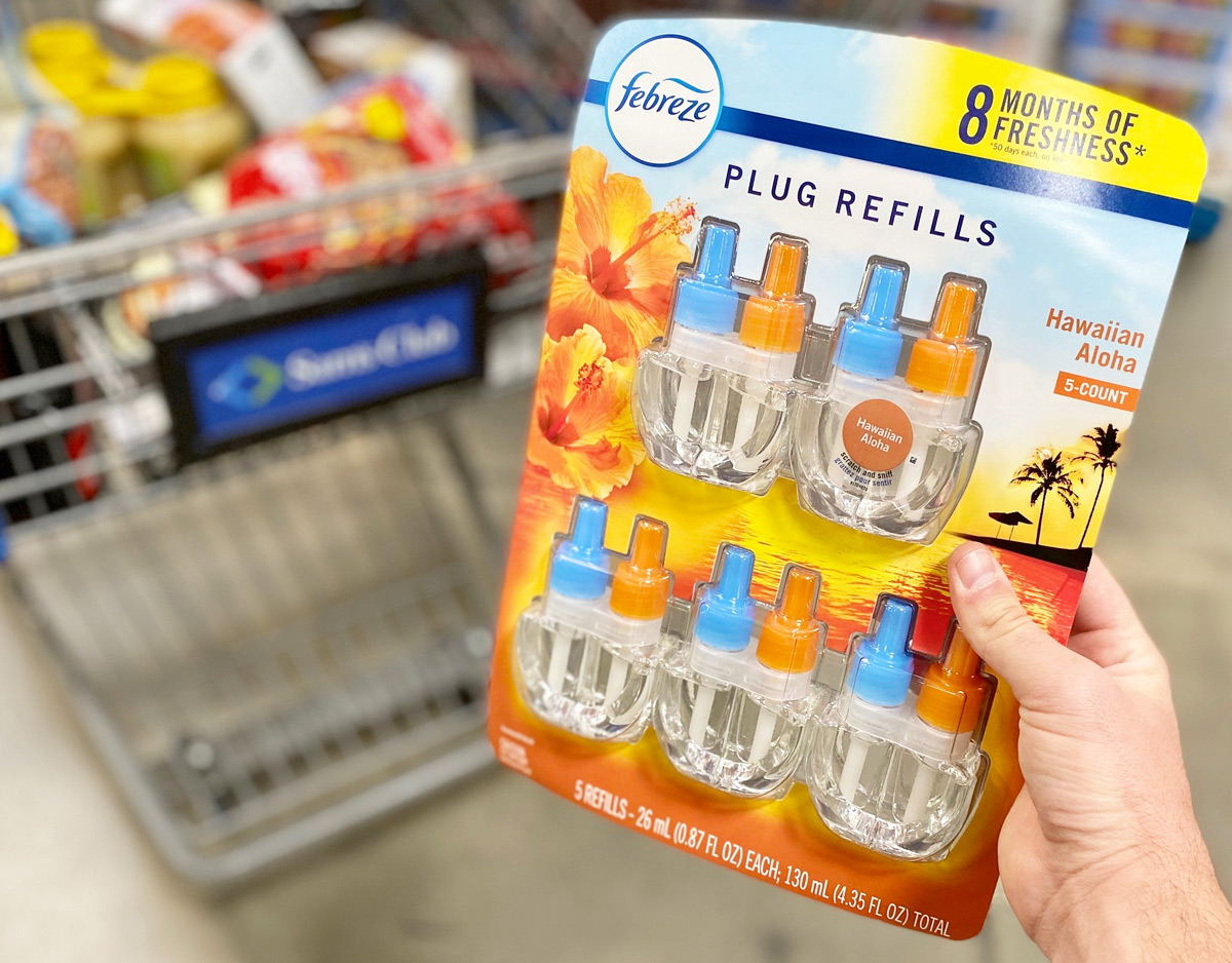 person holding up an orange and blue package of febreze plug refills in hawaiian scent in front of sam's club shopping cart