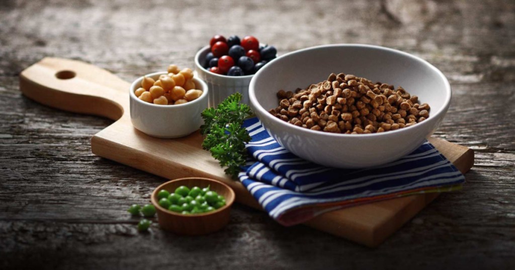 dry cat food in bowl and ingredients in bowls around it on wood cutting board on table