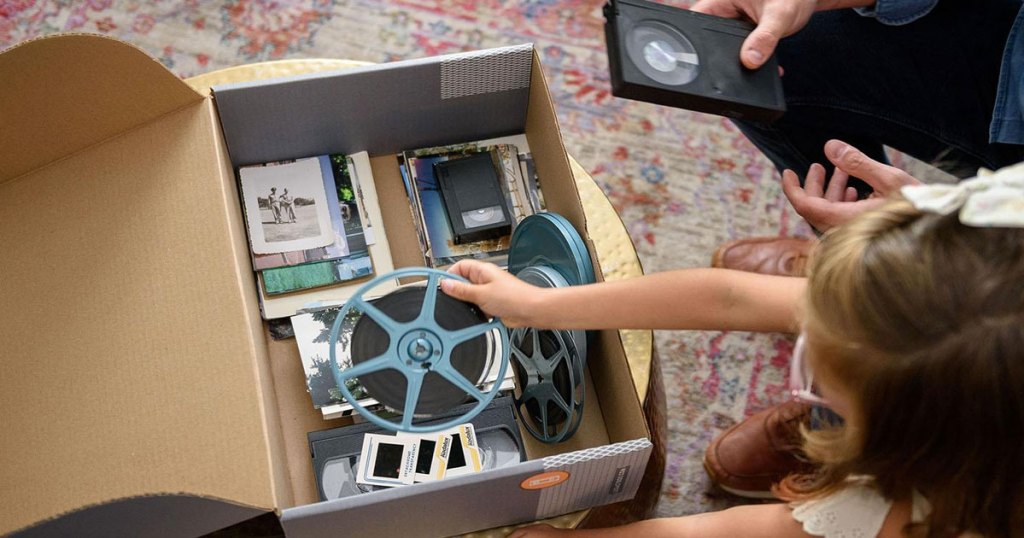 girl with brown hair placing a film reel into a cardboard box full of old photos and vhs tapes