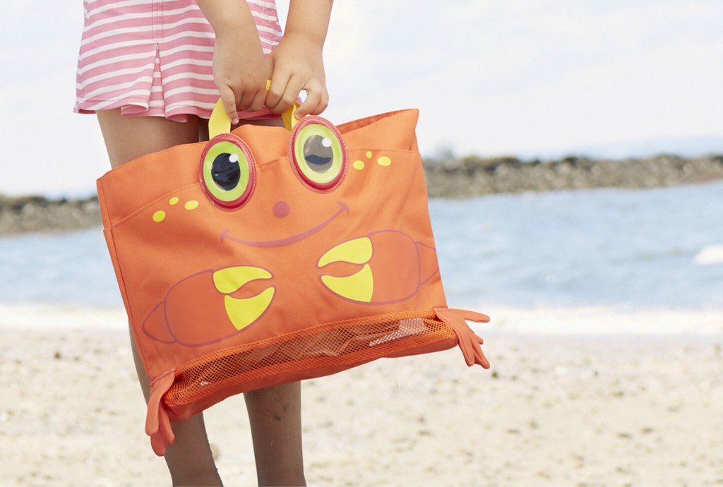 girl standing on beach holding an orange and yellow crab beach bag