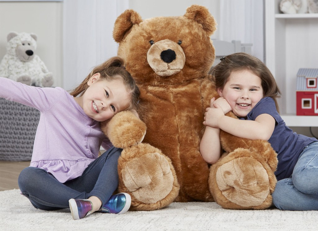 two girls sitting on playroom floor hugging a giant brown teddy bear