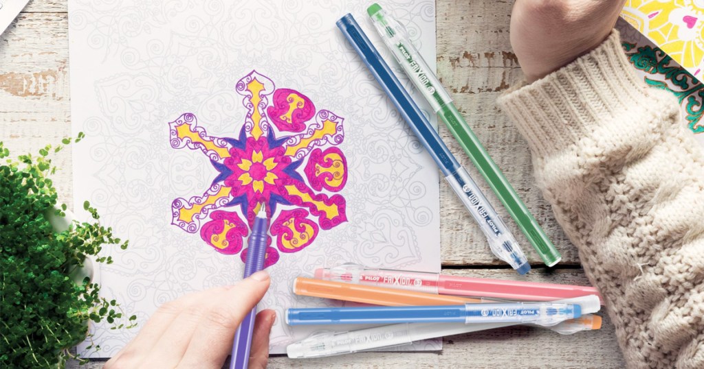 five pens in assorted colors laying on a coloring book page with a person holding a purple pen in their left hand