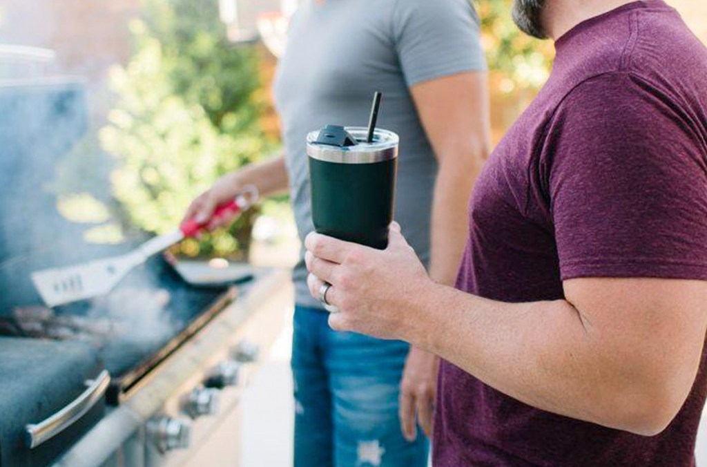 man standing at grill holding a black tumbler in hand with black straw