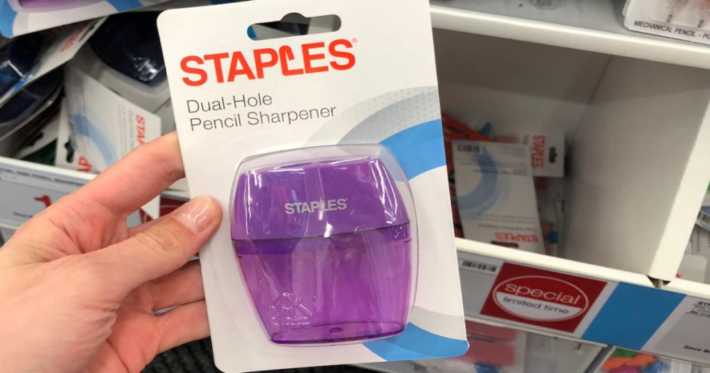 hand holding purple colored Staples Pencil Sharpener. It is purple, in store packaging, held by a hand.
