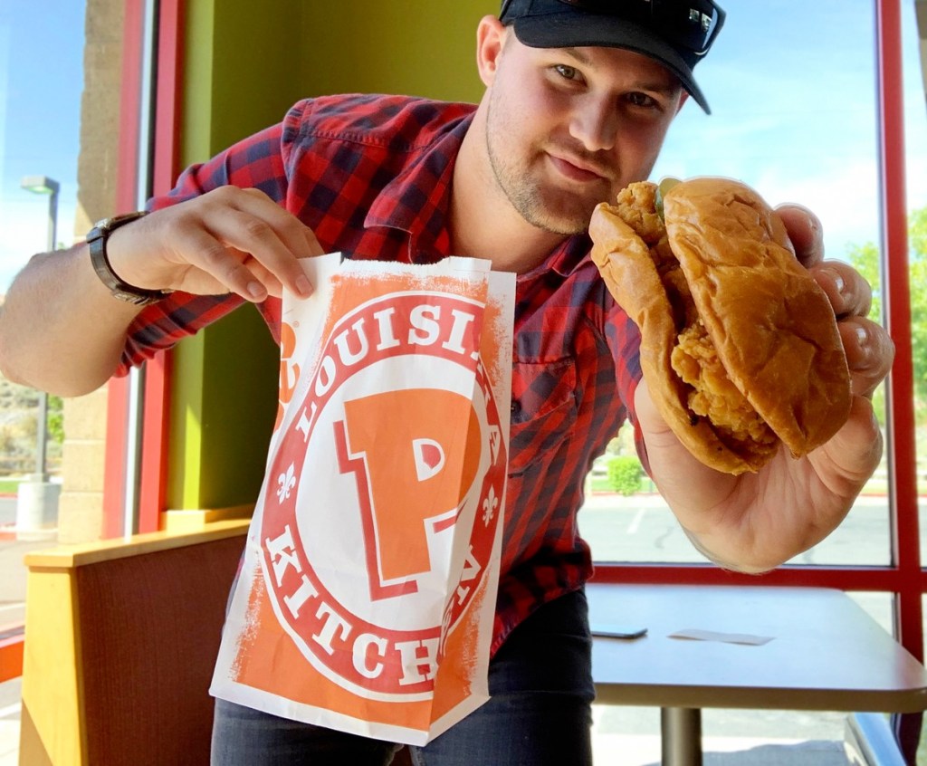 man in red plaid shirt holding popeyes to-go bag in one hand and chicken sandwich in the other