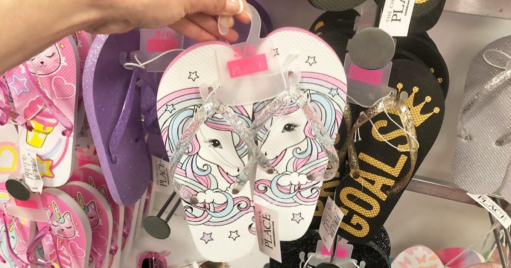 person holding up a pair of girls white unicorn print flip flops with glittery straps