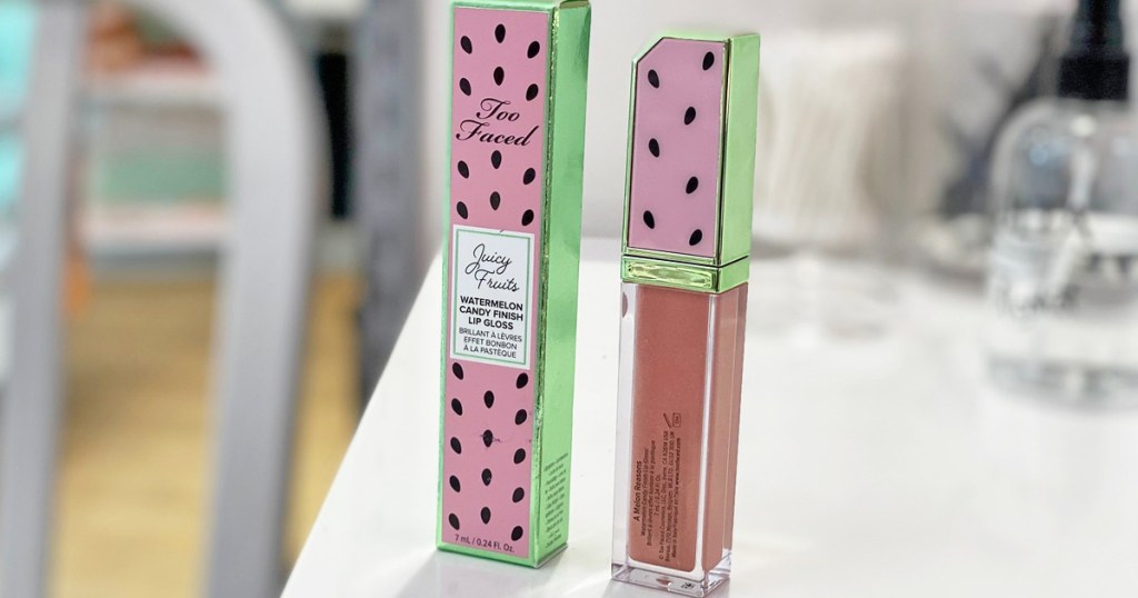 dark pink lip gloss with watermelon print lid standing next to watermelon printed box on a white vanity