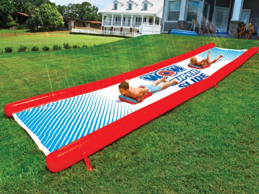 two kids using long water slide on grass in front of home