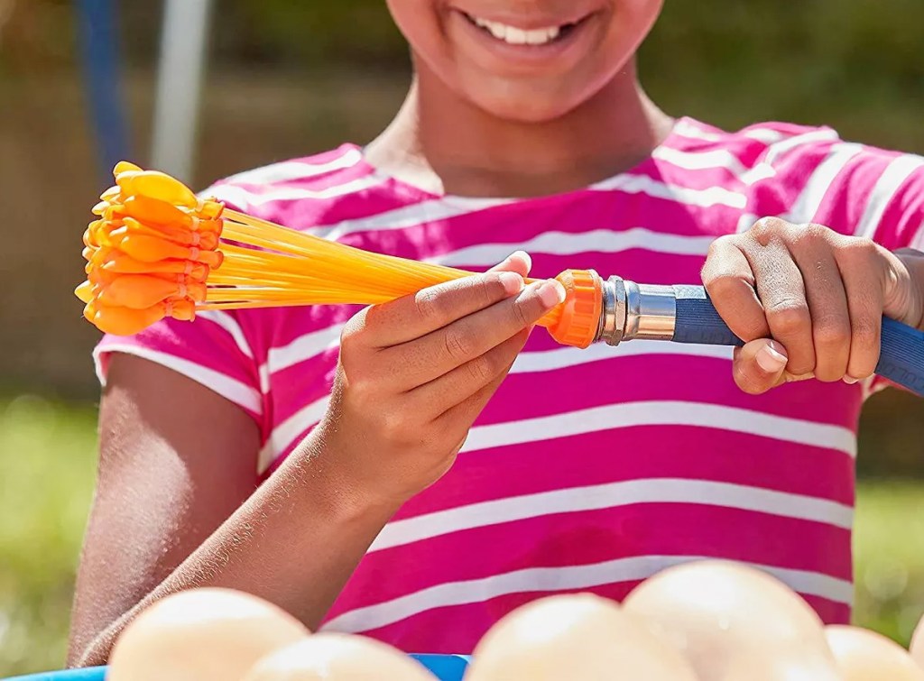 girl in pink stripes shirt attaching orange water balloon filler to garden hose