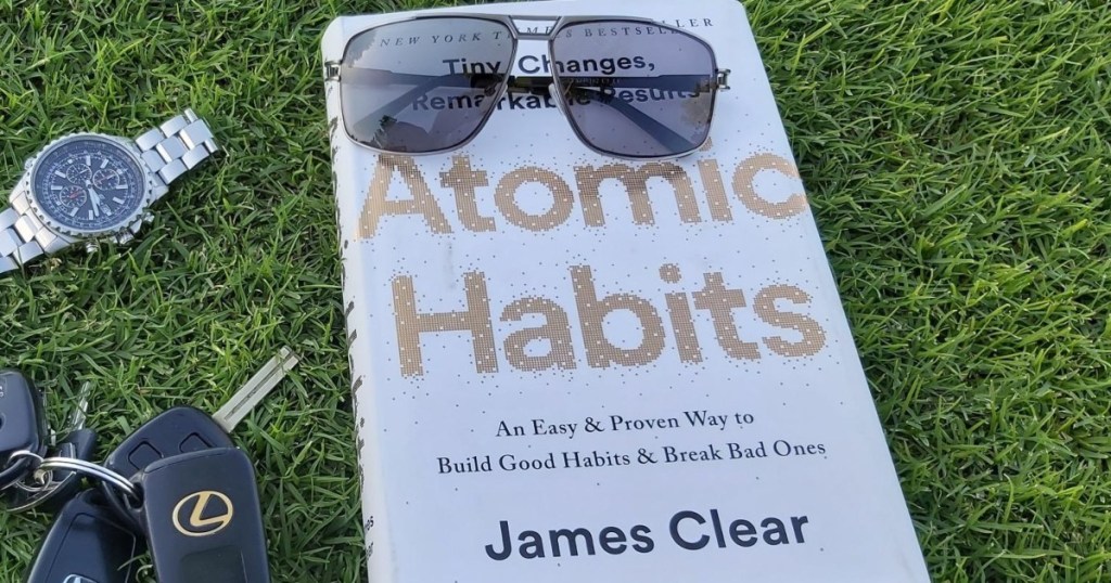 Atomic Habits book in grass with sunglasses, car keys, and watch