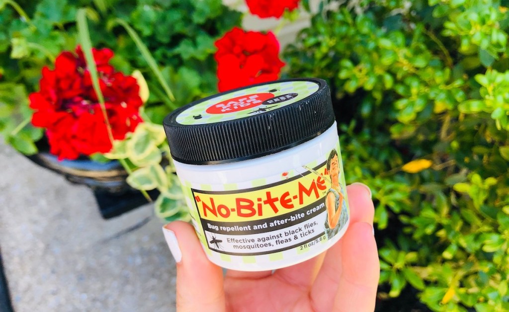 hand holding a tub of no bite me bug repellent in front of plants outside
