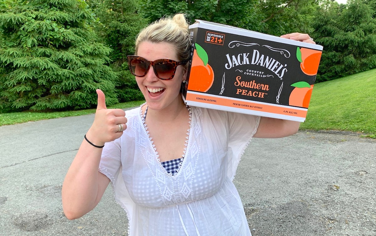 woman holding box of jack daniels refreshers on shoulder giving a thumbs up outside