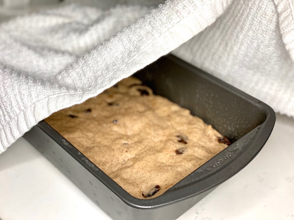 homemade bread dough in loaf pan with towel over the top