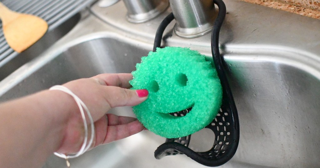 hand holding a green smiley face sponge in stainless steel sink with black holder