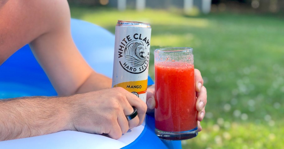 man holding white claw mango can with glass full of red slushie outside in blue blow up pool