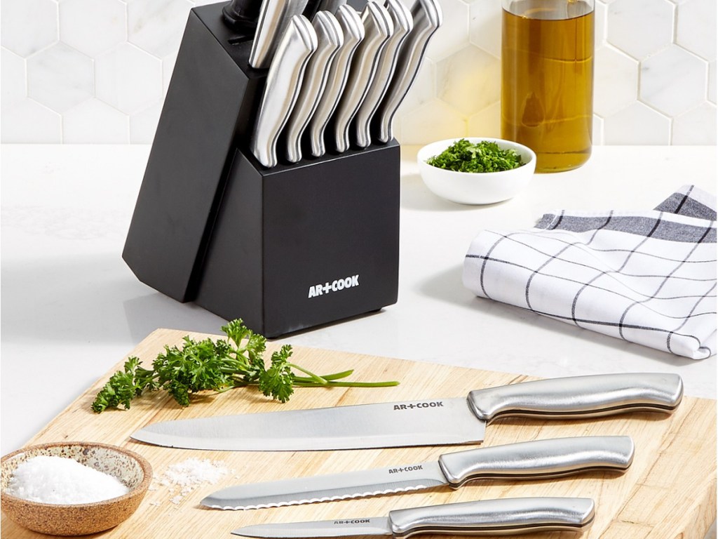 black knife block with stainless steel cutlery on counter with three knives on cutting board and herbs