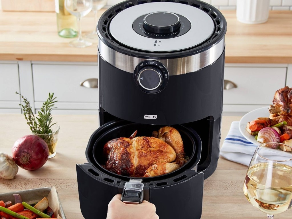 hand opening black air fryer with whole chicken in bottom, on kitchen counter with various food items and wine on counters