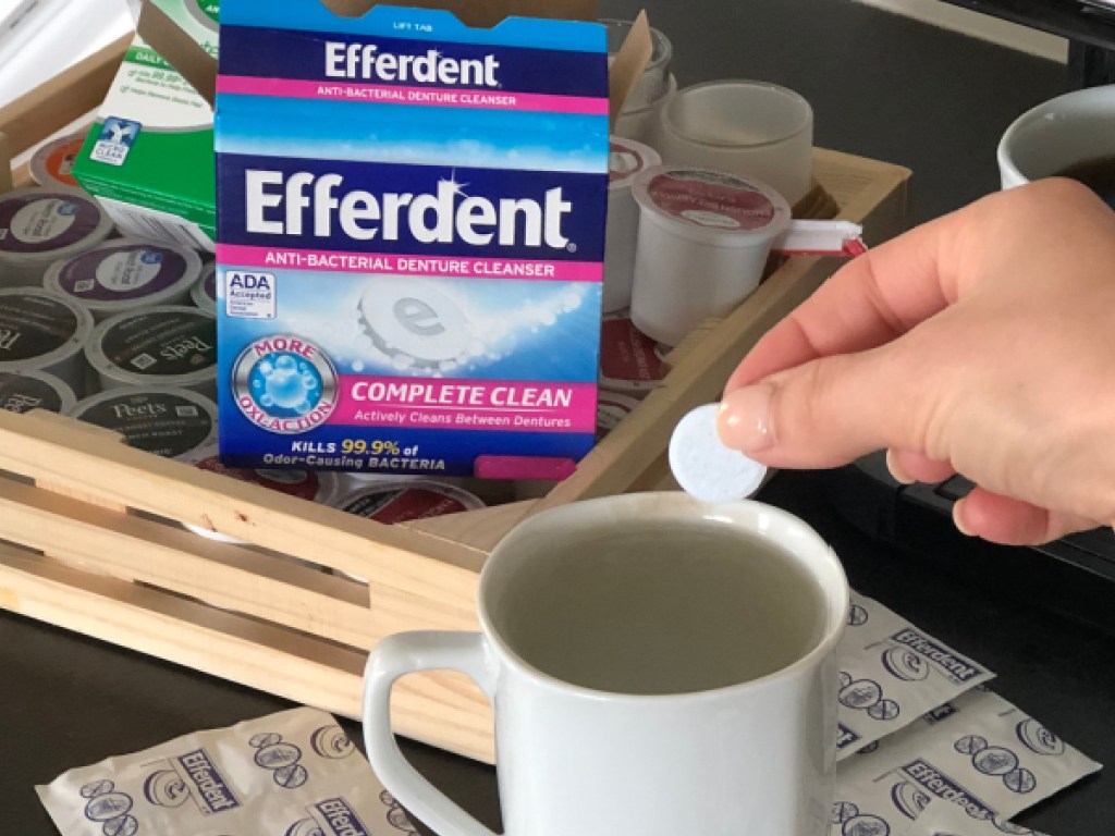 hand putting denture cleaning tablet into white mug with box of denture cleaning tablets and other items on counter in home