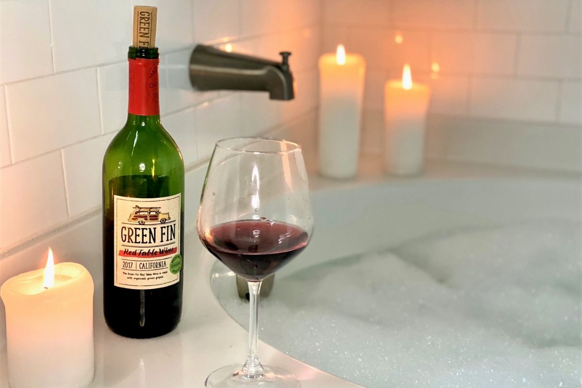 A bottle of red wine and a wine glass next to candles and a bubble bath