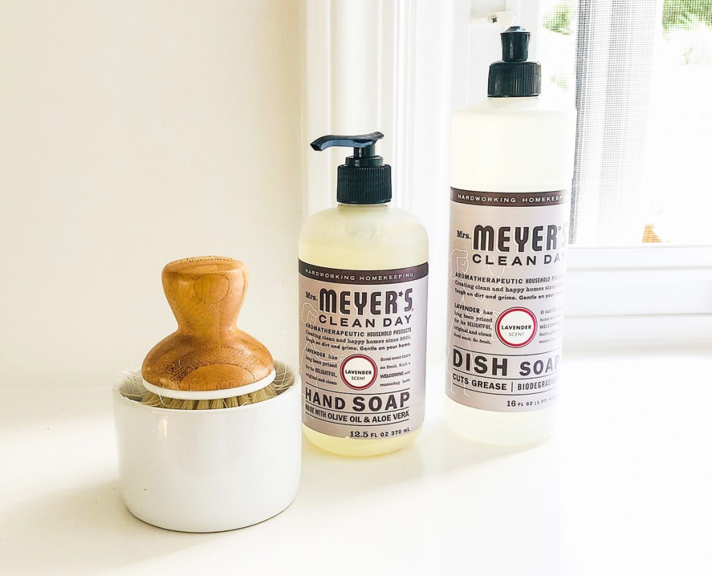two bottles of Mrs Meyers hand soap and dish soap on windowsill next to bamboo dish scrub brush in white holder