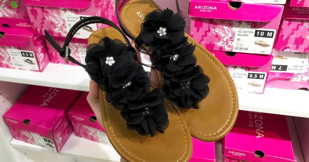 person holding up a pair of sandals with black flowers on them in front of pink shoe boxes