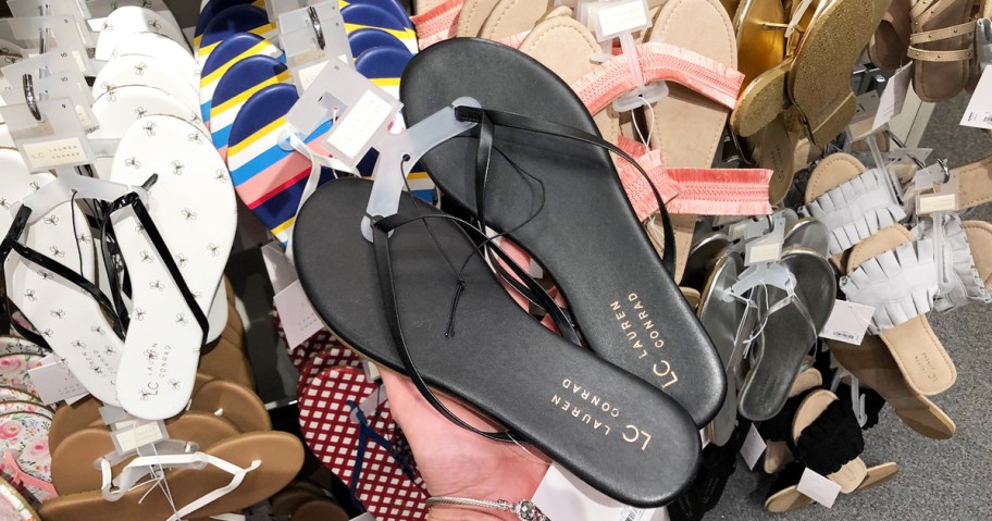 person holding up a black pair of womens flip flops in front of store display rack of sandals
