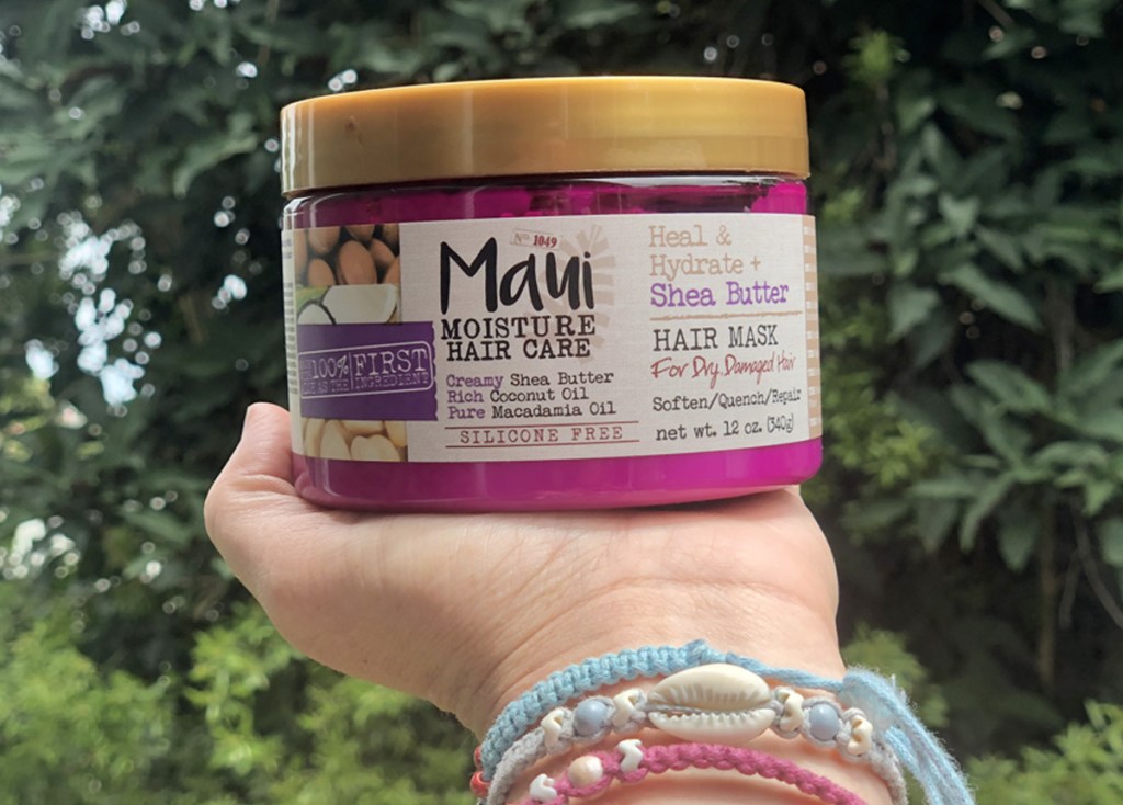 person wearing pink and blue bracelets holding up a purple and gold container of maui moisture hair mask