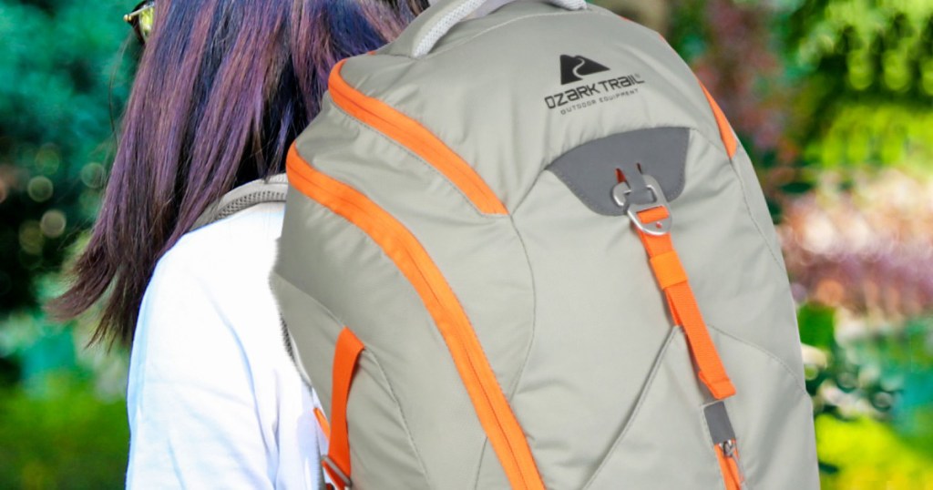woman with brown hair in white sweater wearing a grey backpack with orange zippers