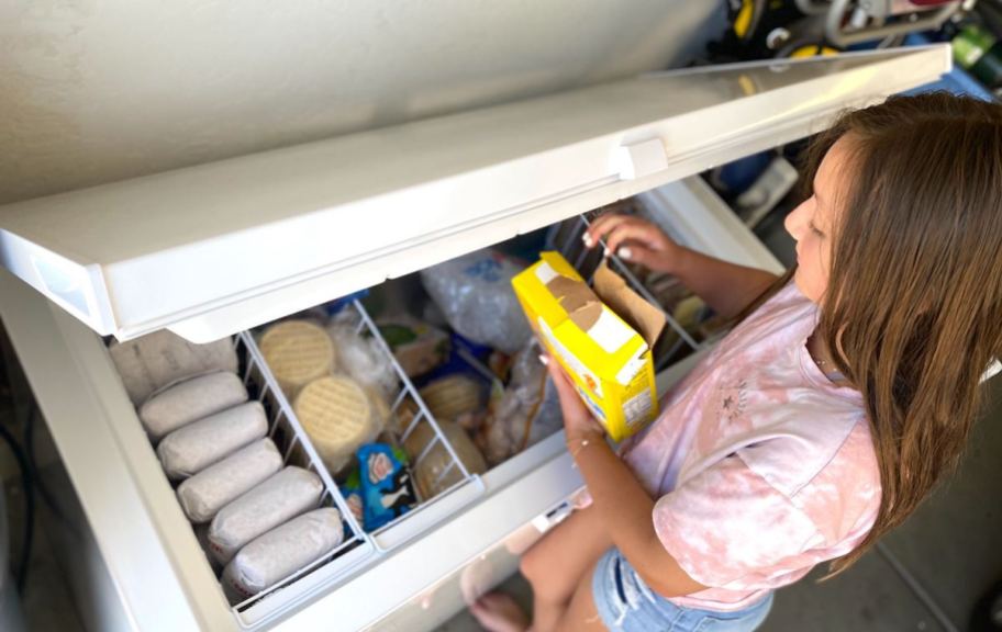 girl holding frozen box of food over stocked open freezer chest