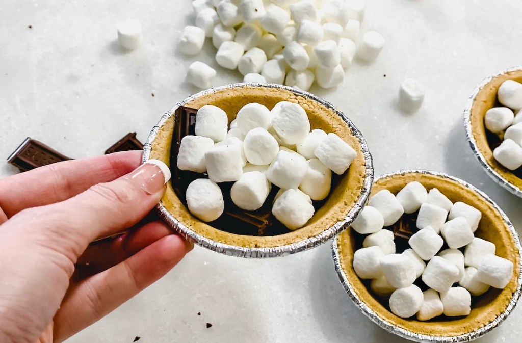 hand holding a smores pie with other ones and marshmallows on counter behind it