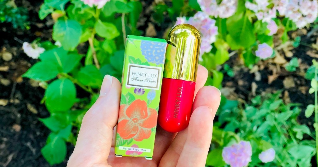 hand holding a hot pink and gold lip balm in front of flower garden