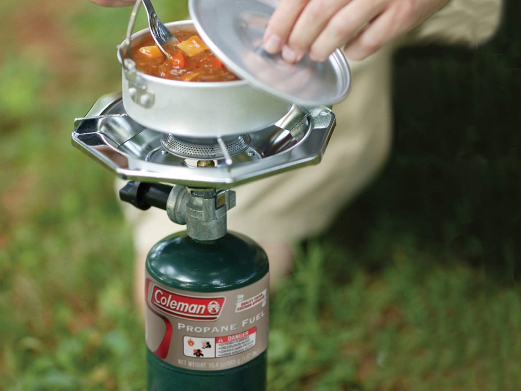 Coleman brand propane stove with a bowl of soup sitting on top and a hand holding the lid