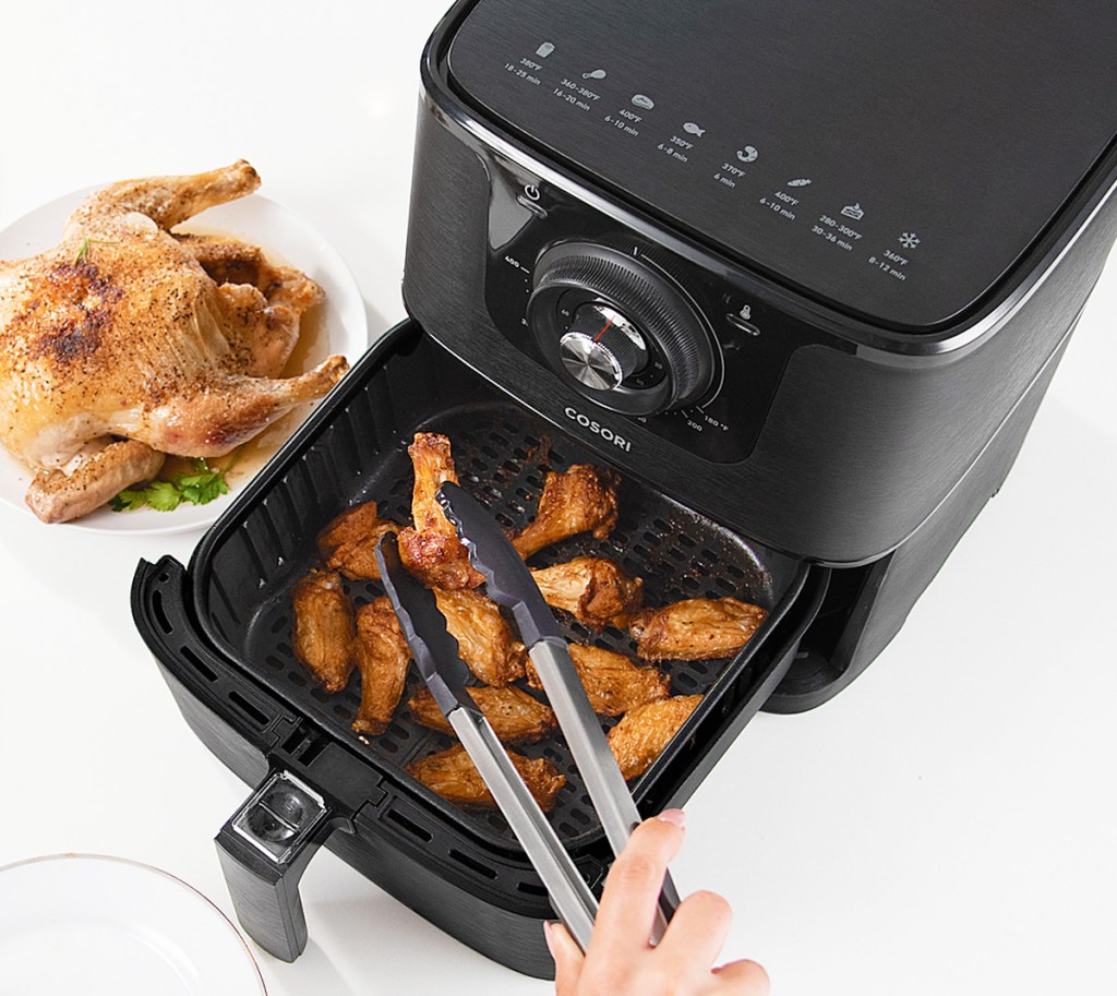 person using tongs to grab chicken wings out of the basket of a black square-shaped air fryer