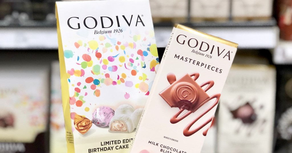 person holding up white bag of Godiva birthday cake chocolates and Godiva milk chocolate bar