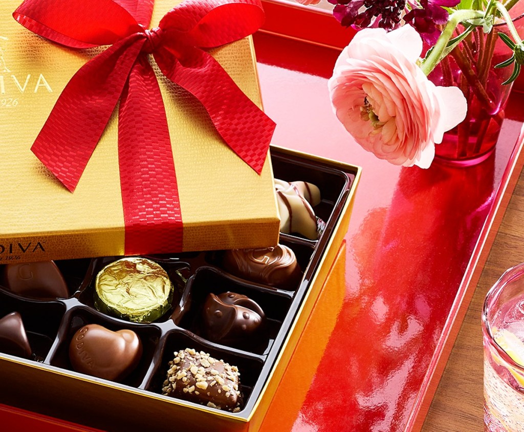 gold box of assorted godiva chocolates with a red ribbon and pink rose next to it