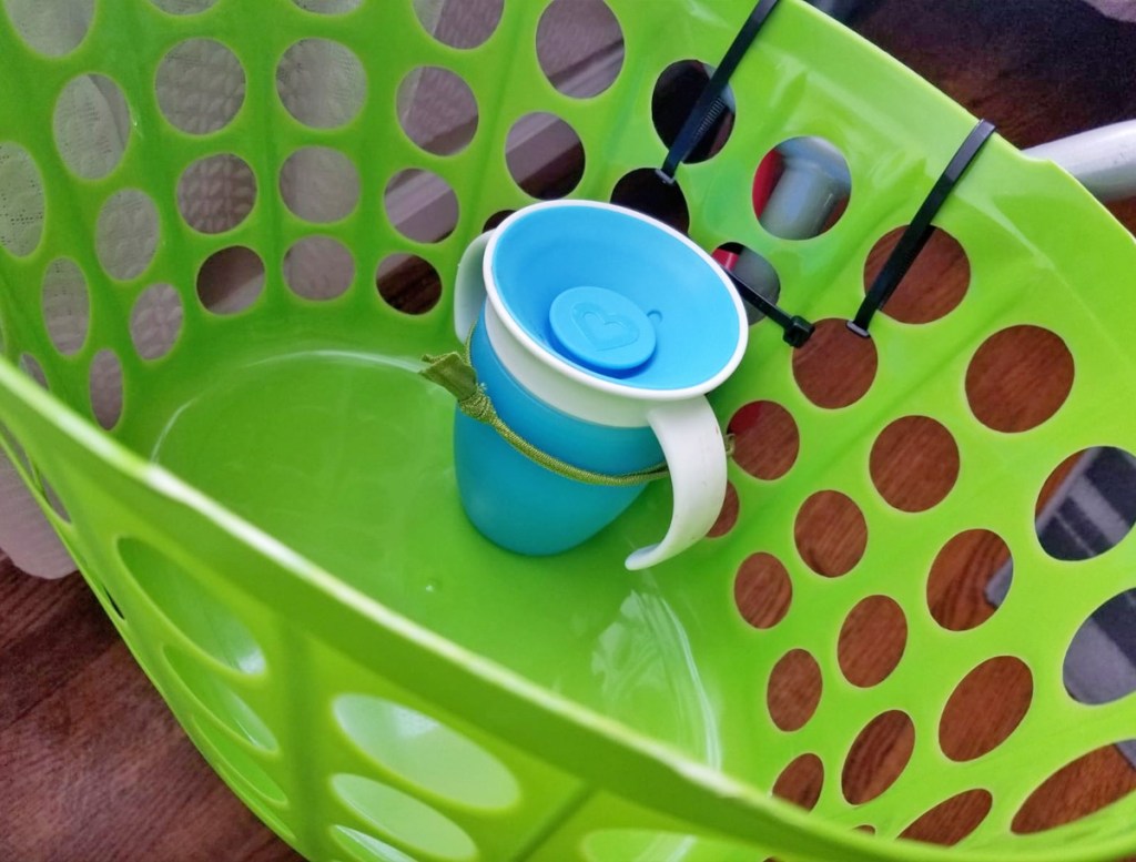 green plastic storage basket attached to front of kids bike with blue sippy cup inside