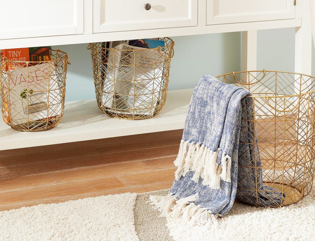 three gold metal wire baskets with two in white console table and one on floor with folded blanket inside