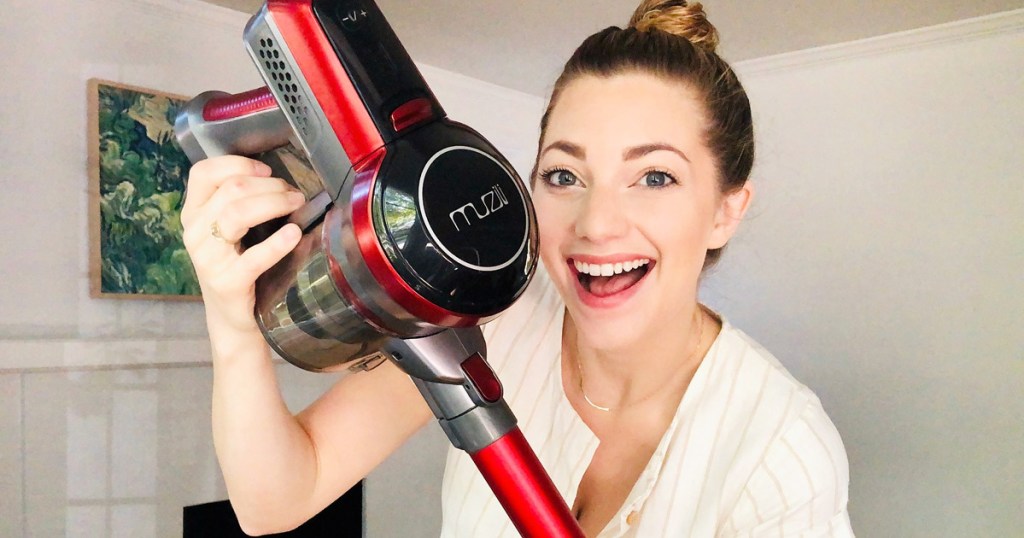 woman with brown hair wearing a white dress holding up a red and grey cordless stick vacuum