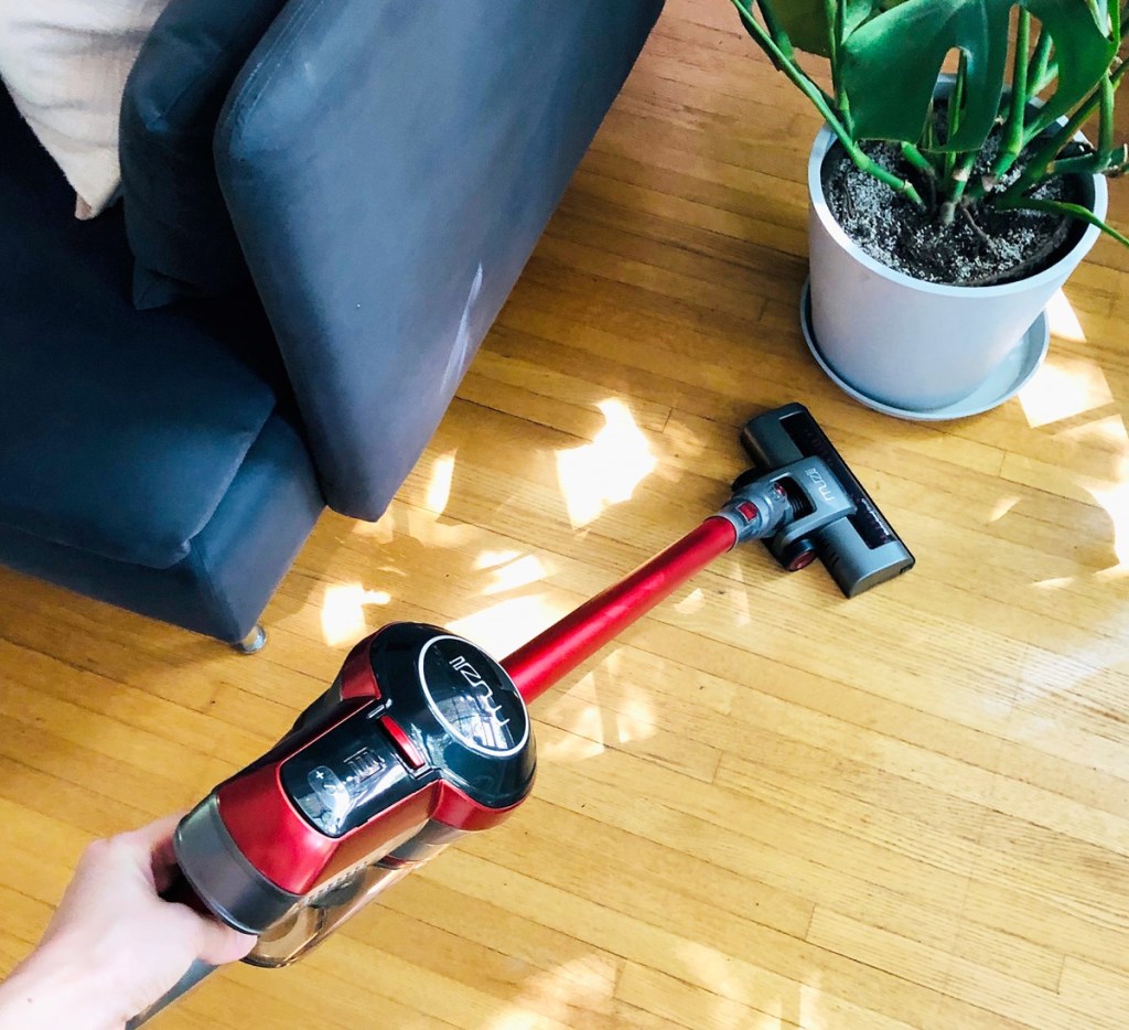 person using a red and grey cordless vacuum on hardwood floor near a blue couch and potted plant