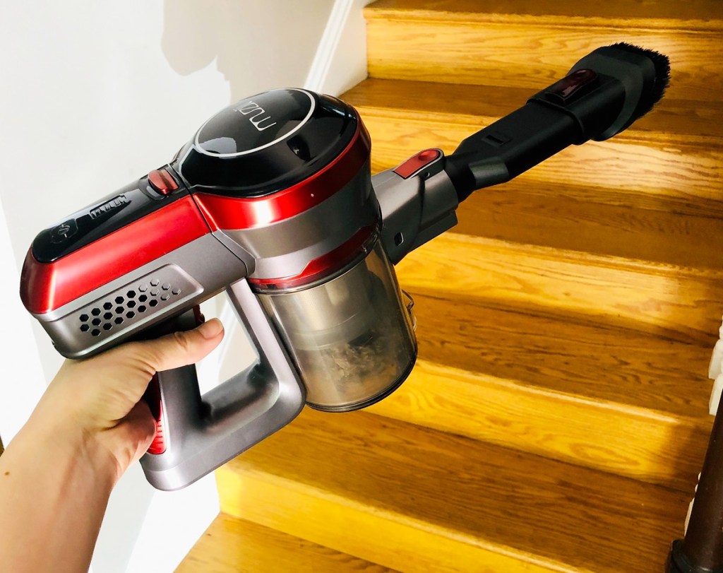 woman holding a red and grey cordless vacuum cleaner with brush attachment near wood staircase