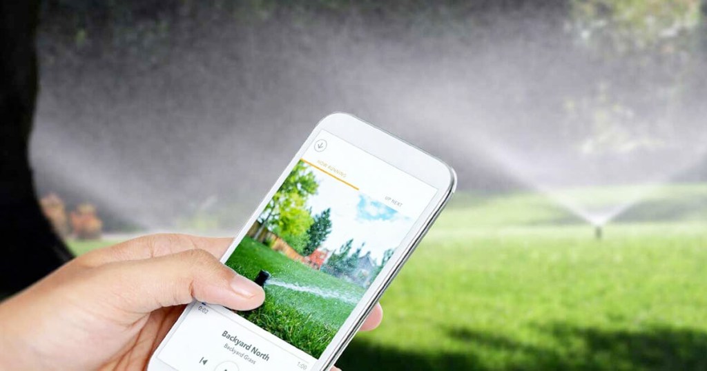 person holding white smartphone with rachio app controlling a lawn sprinkler in the background