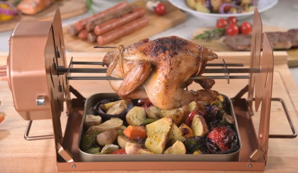 A rotisserie cooking machine with a chicken and grilled veggies cooking