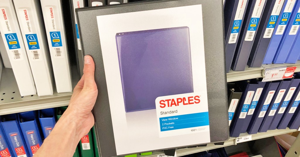 person holding up a black Staples brand 1" binder in front of display of other binders
