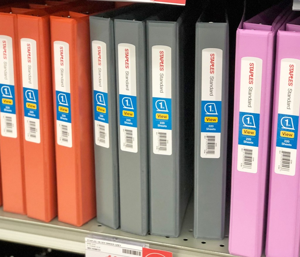 Staples brand 1" binders in orange, grey, and pink colors on store shelf