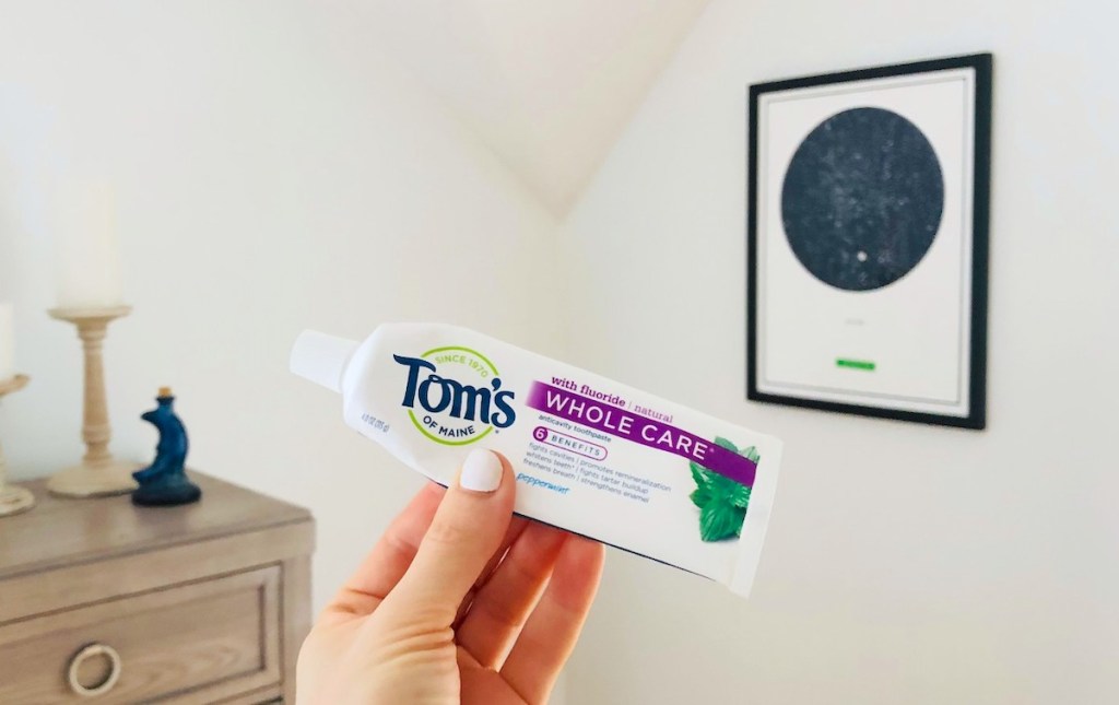hand holding a tube of toms toothpaste in front of hanging picture