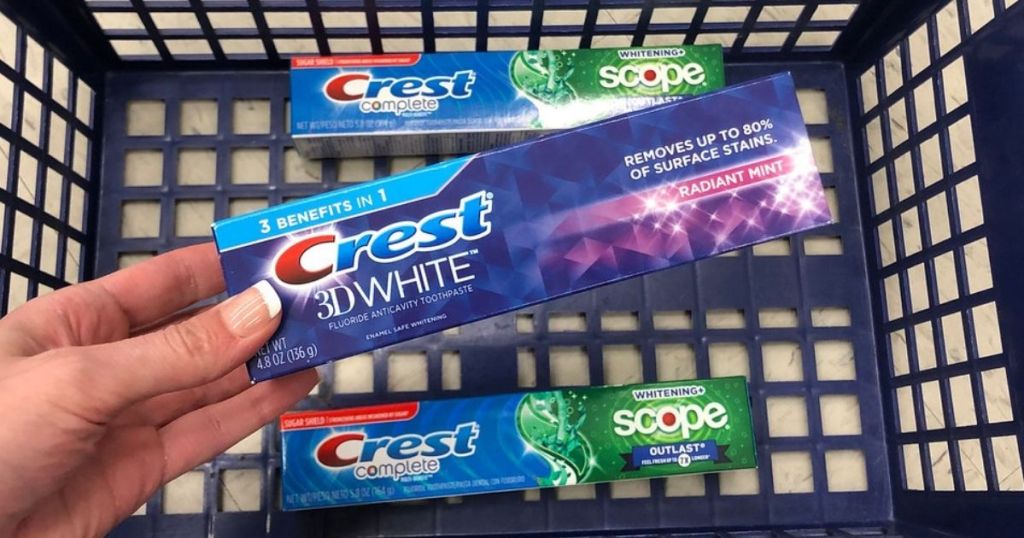 woman's hand holding tube of crest toothpaste over shopping basket with two more boxes of crest toothpaste in it